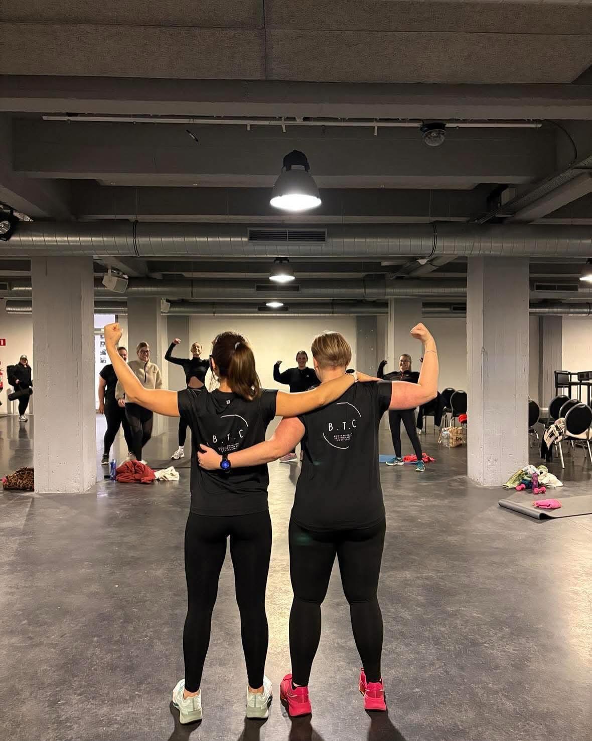 Deux coachs BTC vues de dos, bras levés, avec le groupe en arrière-plan pendant un cours collectif en salle